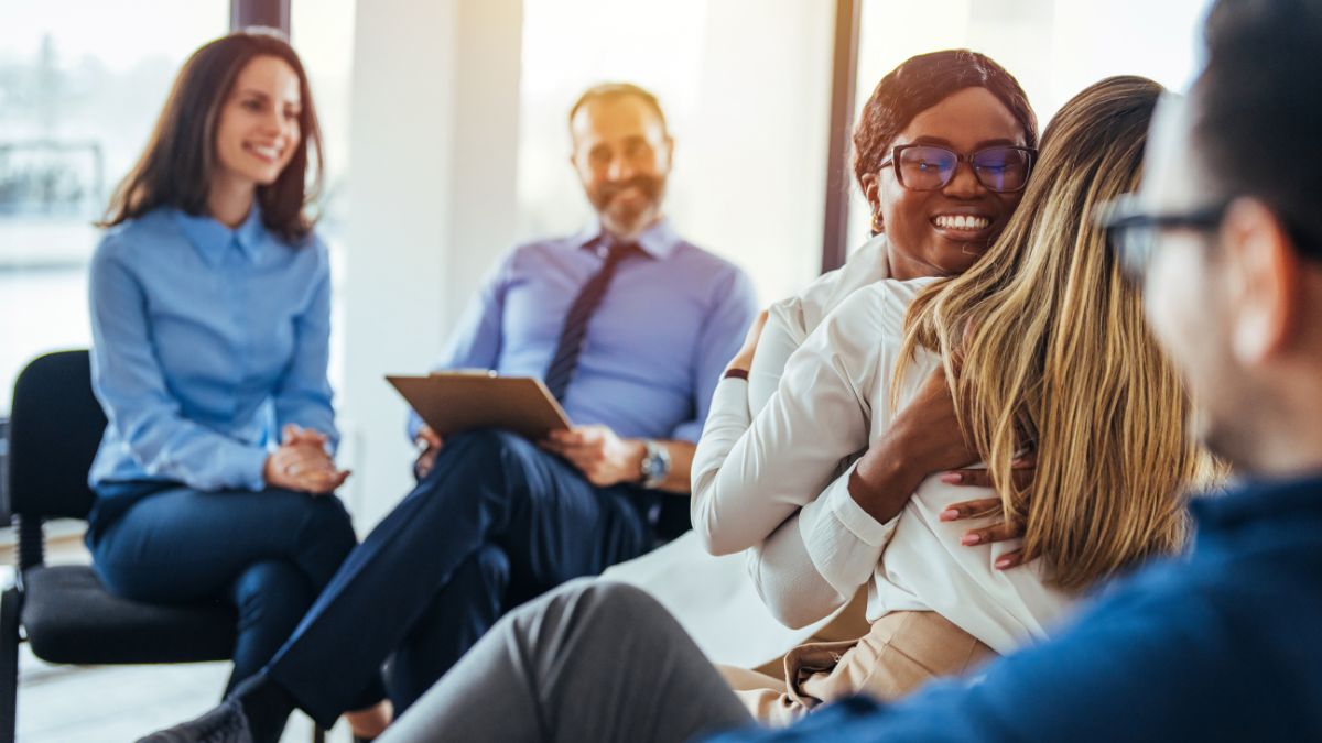 A photo of two employees hugging while in a group meeting with the other employees